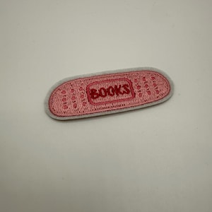 Könnte beinhalten: Ein rosa bestickter Aufnäher mit dem Wort „BOOKS“ in roten Buchstaben. Der Aufnäher hat eine rechteckige Form mit abgerundeten Kanten und einem hellgrauen Rand. Er ist für Kleidung oder Accessoires bestimmt.