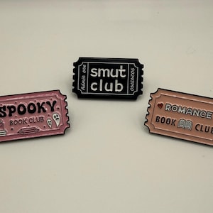 Pode incluir: Três pinos de esmalte em forma de bilhetes vintage. Um pino rosa diz "Spooky Book Club" com um fantasma e um gráfico de cristal. Um pino preto diz "smut club". Um pino rosa diz "Romance Book Club" com um coração e um gráfico de livro.