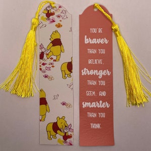 Puede incluir: Dos marcapáginas decorativos con borlas amarillas. Uno presenta ilustraciones de Winnie the Pooh y Piglet sobre un fondo blanco. El otro es de color coral con el texto "You're braver than you believe, stronger than you seem, and smarter than you think."