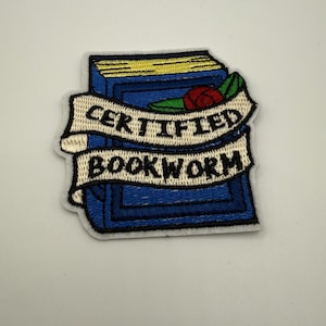 May include: Embroidered patch depicting a blue book with gold pages and a red rose. A banner across the book reads "CERTIFIED BOOKWORM" in black text. The patch has a white border, suitable for application on clothing or accessories.