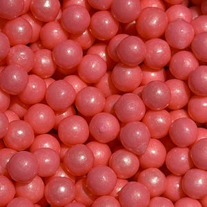 May include: Close-up of a pile of pink, round candies. The candies have a shiny, pearl-like finish and are densely packed together. The color is a vibrant, rosy pink, and the overall effect is visually appealing.