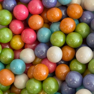 May include: A close-up shot of a collection of colorful, round candies. The candies are in various colors, including pink, orange, green, blue, purple, and white. The candies have a shiny, pearl-like finish.