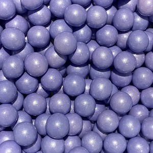 May include: A close-up shot of a pile of lavender-colored, spherical candies. Each candy has a smooth, shiny surface with subtle highlights. The candies are densely packed together, filling the frame.