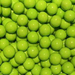 May include: A close-up shot of a pile of bright green, round candies. The candies have a glossy finish and are evenly sized, filling the frame. The overall effect is a vibrant, textured display of confectionery.