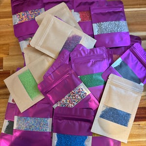 May include: A collection of purple and brown pouches filled with colorful sprinkles. The pouches have a clear window to display the contents. The sprinkles are in various colors and sizes, perfect for decorating baked goods.