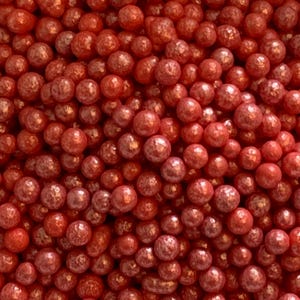 May include: Close-up of a collection of small, round, red-colored sprinkles. The sprinkles have a shiny, pearl-like finish, and are densely packed together, creating a textured surface. These are likely used for decorating baked goods.