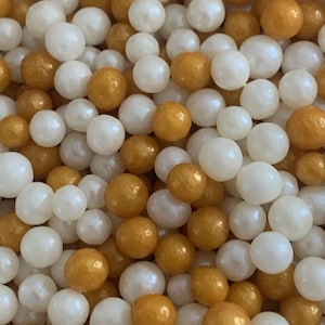 May include: Close-up of a collection of round sprinkles in white and gold colors. The sprinkles are small and spherical, creating a textured, detailed surface. These are likely used for decorating baked goods.
