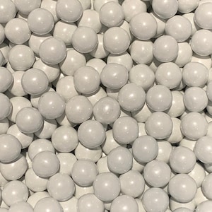 May include: An overhead shot of a pile of small, round, light gray candies. The candies are tightly packed together, filling the frame. The candies have a smooth, glossy finish, and the lighting creates highlights and shadows.