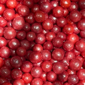 May include: Close-up of a pile of red, round candies. The candies have a shiny, slightly textured surface and vary in size. The image is filled with the candies, creating a dense, textured pattern. The color is a deep, vibrant red.