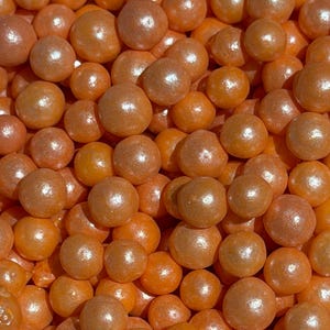 May include: Close-up of a pile of orange, spherical sprinkles with a pearlescent sheen. The sprinkles are densely packed, creating a textured surface. The color is a warm, inviting orange.