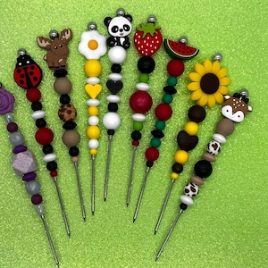 Peut inclure: Une collection de brochettes décoratives avec différents motifs. Les motifs comprennent une coccinelle, un orignal, un panda, une fraise, une pastèque et un tournesol. Les brochettes sont en métal et ont des perles colorées.