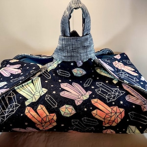 May include: A large tote bag featuring a dark blue fabric adorned with a pattern of colourful crystals and geometric shapes. The bag has a handle made from a blue and white plaid fabric.