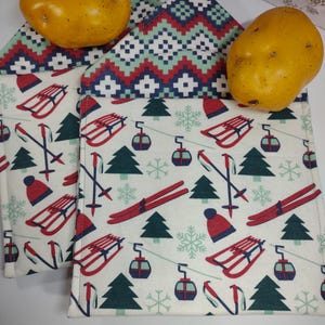 May include: Two white fabric napkins with a winter sports pattern featuring red sleds, skis, hats, and green trees. The napkins have a patterned border in red, white, and teal. Two yellow potatoes are on top.