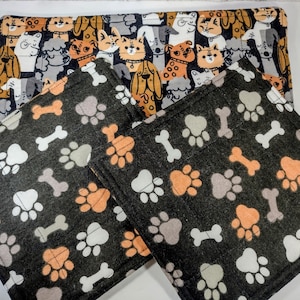 May include: A set of pet-themed items, including a rectangular mat and three square coasters. The mat features a pattern of various dog and cat illustrations in orange, black, white, and gray. The coasters have a black background with paw prints and bone designs in orange, gray, and white.