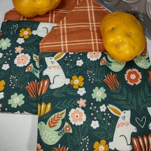 Può includere: Due presine in tessuto verde con un coniglio bianco e un motivo floreale. Le presine hanno un retro a quadri marroni e bianchi.