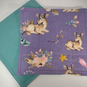 Puede incluir: Dos cuadrados de tela. Uno es turquesa, acolchado con costuras diagonales. El otro es morado, con un estampado de ciervos, flores y plumas. Los ciervos son de color beige y blanco, con tocados florales. Las flores son rosas y crema.