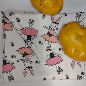 May include: White fabric envelope with a repeating pattern of pink ballerina bunnies. The envelope has a flap with a pink stitched edge. Two yellow potatoes are in the background.
