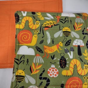 Puede incluir: Dos artículos de tela rectangulares. Uno es de color naranja liso con líneas de cuadrícula cosidas. El otro es verde con un patrón repetido de insectos, caracoles y setas de colores. El artículo estampado está superpuesto al artículo naranja.