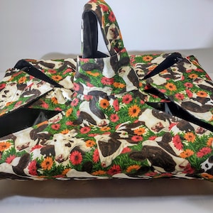 May include: A large tote bag with a cow and floral print. The bag features a repeating pattern of black and white cows, red and orange flowers, and green foliage. The bag has a black interior and a matching handle.