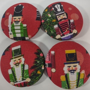 May include: Four round Christmas-themed fabric buttons with red backgrounds. Each button features a different illustration of a nutcracker soldier. Some buttons show a Christmas tree. The nutcrackers are in green, red, and white.