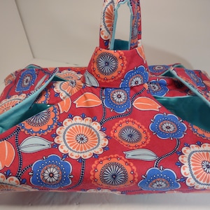 May include: A red fabric pet carrier with a floral pattern in blue and orange. The carrier has a turquoise interior and trim, with a matching handle. The design features large stylized flowers and leaves.