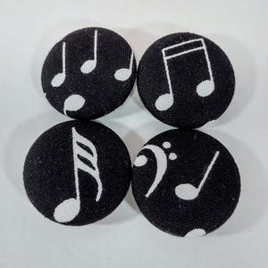 以下が含まれることがあります： 白の音符が付いた黒い布で覆われた4つのボタン。 各ボタンには、四分音符、八分音符、低音部記号など、さまざまな音楽記号が表示されています。 ボタンは丸く、質感があります。