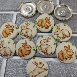Puede incluir: Un conjunto de botones redondos cubiertos de tela con ilustraciones de conejos. Los botones, de unos 2,5 cm de diámetro, tienen un fondo crema con detalles verdes. Los diseños de conejos varían, y algunos muestran los respaldos metálicos.