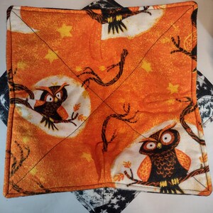 May include: Square orange fabric pot holder with a Halloween owl design. The fabric features owls perched on branches against a full moon and star background. The pot holder is trimmed with black stitching.
