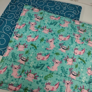 May include: Two square quilted pot holders. One is teal with a white swirl pattern. The other is turquoise with a pattern of pink axolotls, green leaves, and bubbles. The pot holders are stitched with black thread.