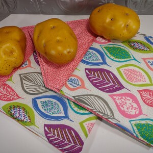 May include: Three yellow potatoes sit on two decorative fabric placemats. The placemats have a white background with colourful leaf patterns in pink, purple, green, and blue. The placemats are edged with a pink floral pattern.