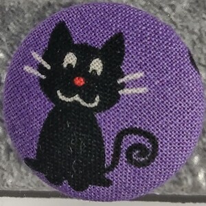 Può includere: Un bottone rotondo con sfondo viola e un'illustrazione di un gatto nero. Il gatto ha baffi bianchi, un naso rosso e una bocca sorridente. Il bottone è probabilmente fatto di tessuto ed è un oggetto decorativo.