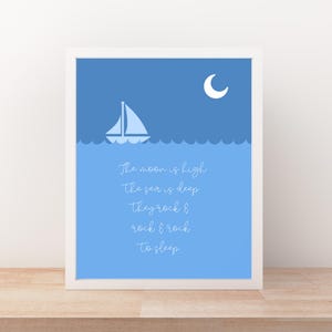 Puede incluir: Una impresión enmarcada con un tema náutico. La obra de arte presenta un velero blanco sobre agua azul bajo una luna creciente. El texto "The moon is high, the sea is deep, they rock & rock & rock to sleep" está escrito en cursiva.