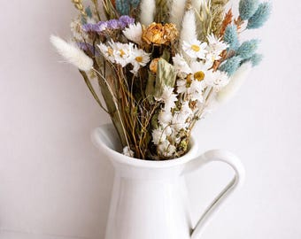 Op de afbeelding: Een witte keramische kan bevat een boeket van gedroogde bloemen. Het boeket bevat witte madeliefjes, blauwe, oranje en paarse accenten en diverse gedroogde grassen. De kan heeft een handvat en staat op een wit oppervlak.