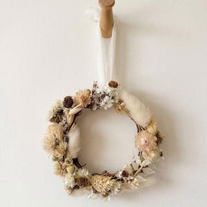 May include: A circular wreath made of dried flowers and grasses in shades of beige, cream, and brown. The wreath is suspended by a white ribbon from a wooden peg on a white wall. The wreath features various textures and shapes.
