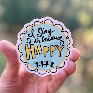 May include: A round sticker with a blue background and white scalloped border. The sticker says "I sing because I'm happy" in black and yellow lettering. There are five small black birds singing on a branch below the text.