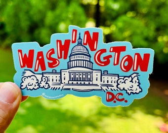 Pode incluir: Um adesivo azul e vermelho com o texto "I'm in Washington D.C." e uma ilustração branca do Capitólio.