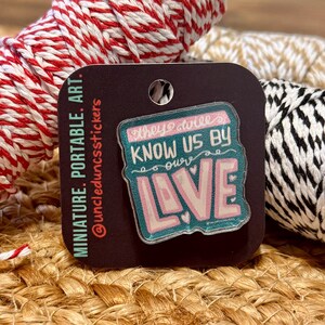 May include: A rectangular pin with the text "They will know us by our love" in pink and teal. The pin is on a dark brown card with the words "miniature. portable. art." and the seller's Instagram handle.