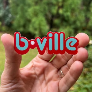 May include: A red and blue sticker with the text "b-ville" in a rounded font.
