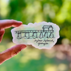 May include: A hand-drawn illustration of a pier with a small building on the end. The pier is over water and the text "tybee island, georgia" is written below the pier.