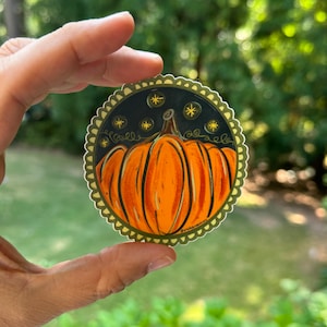May include: A round sticker with a dark green border and a white scalloped edge. The sticker features a hand-painted illustration of an orange pumpkin with a dark green stem and vines. The background is black with gold stars.