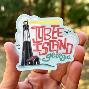 May include: A sticker featuring a black and white lighthouse with a red roof, the words "Tybee Island Georgia" in red and green, and two small birds. The sticker is held in a hand.