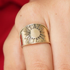 Könnte beinhalten: Ein goldfarbener Ring mit einem Sonnenmuster. Der Ring hat ein breites Band mit einer detaillierten Sonnengrafik in der Mitte. Der Ring wird am Finger getragen.