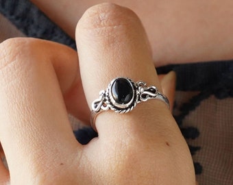 Könnte beinhalten: Ein Silberring mit einem zentralen ovalen schwarzen Stein, gefasst in ein dekoratives Metallband mit gedrehten Kordeldetails und komplizierten Schnörkelmustern an den Seiten, getragen an einem Finger vor einem dunklen Stoff mit schwarzer Stickerei.