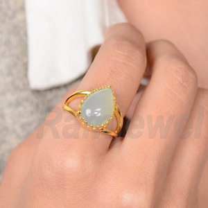 May include: A gold ring with a pale blue, pear-shaped gemstone. The ring has an openwork design and a split shank. The ring is worn on a finger. The gemstone is set in a gold bezel.