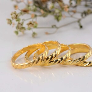 Puede incluir: Cuatro anillos dorados con un diseño de cuerda trenzada. Los anillos se muestran sobre una superficie blanca, con un fondo borroso de flores secas. Los anillos están hechos de un material metálico brillante.