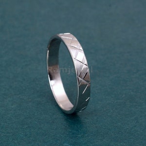 Puede incluir: Un anillo plateado con un patrón geométrico de líneas que se cruzan. El anillo se encuentra sobre un fondo verde azulado, resaltando su superficie pulida y su intrincado diseño. La banda del anillo tiene un patrón texturizado en forma de diamante.