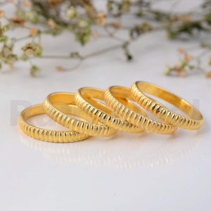Peut inclure: Quatre bagues dorées avec un motif texturé et strié. Les bagues sont disposées en ligne légèrement incurvée, avec un arrière-plan flou de blanc et de fleurs séchées. Les bagues sont en métal brillant.
