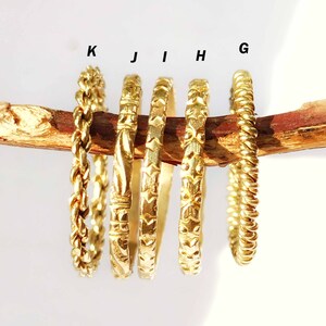 May include: A collection of gold-colored rings with various textures and designs, including braided, patterned, and textured surfaces. The rings are displayed on a brown branch against a white background. Each ring is labeled with a letter.