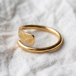 Könnte beinhalten: Ein goldfarbener offener Ring mit einer glatten, abgerundeten Ringschiene, die in ein flaches, strukturiertes Ende mit gehämmertem Finish übergeht. Die Metalloberfläche reflektiert das Licht und betont den Kontrast zwischen dem polierten Band.