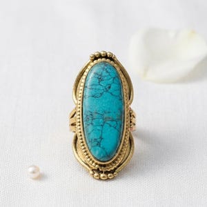 Op de afbeelding: Een goudkleurige ring met een grote, ovale turkooiskleurige steen met donkere adering. De ring heeft een sierlijke, gedetailleerde zetting met een kralenrand en decoratieve accenten. De achtergrond is een witte, getextureerde ondergrond.